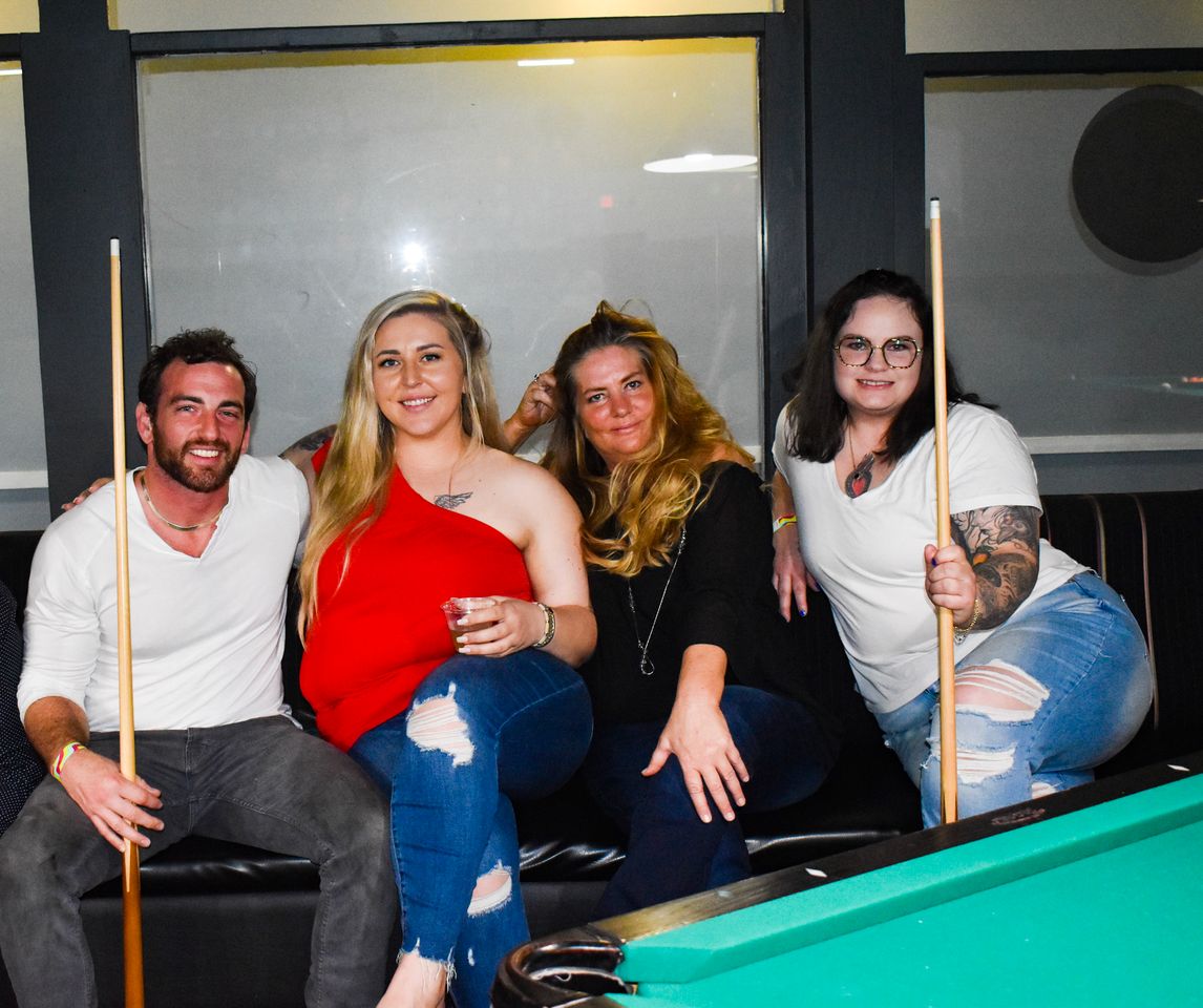 A group of four friends holding pool cues inside of the Rigby's Entertainment Complex Billiards Room